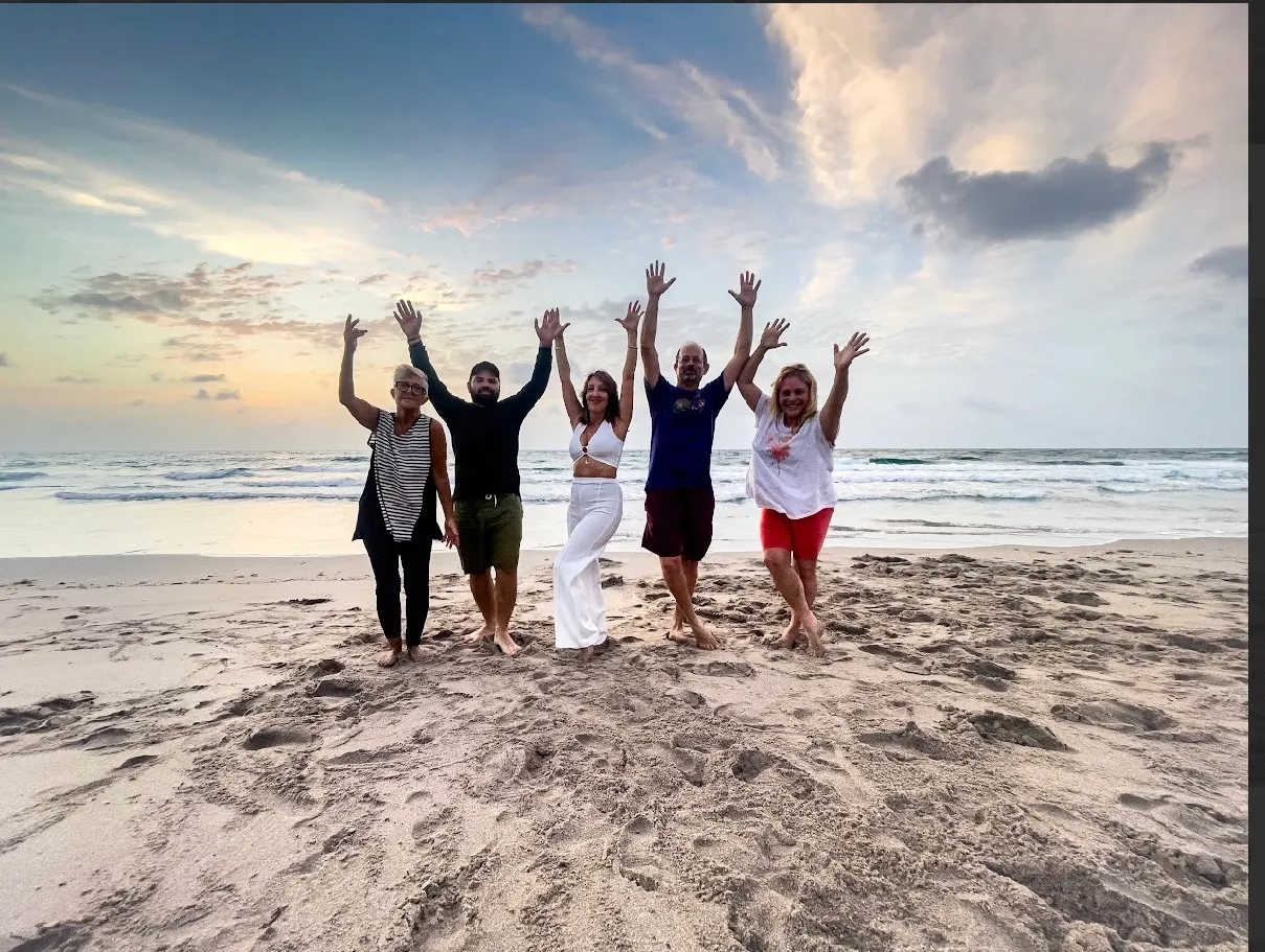 Five people standing barefoot on a sandy beach with arms raised, ocean waves and a cloudy sky at sunset in the background.