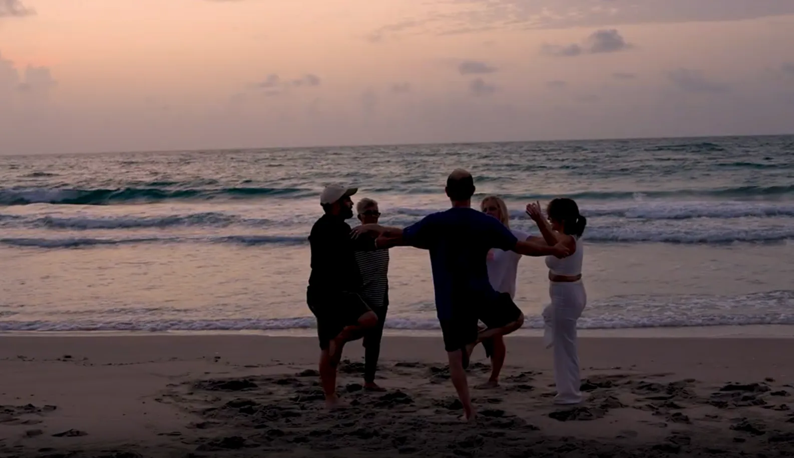 Group of five people standing on one leg holding hands on a beach at sunset with ocean waves in the background.