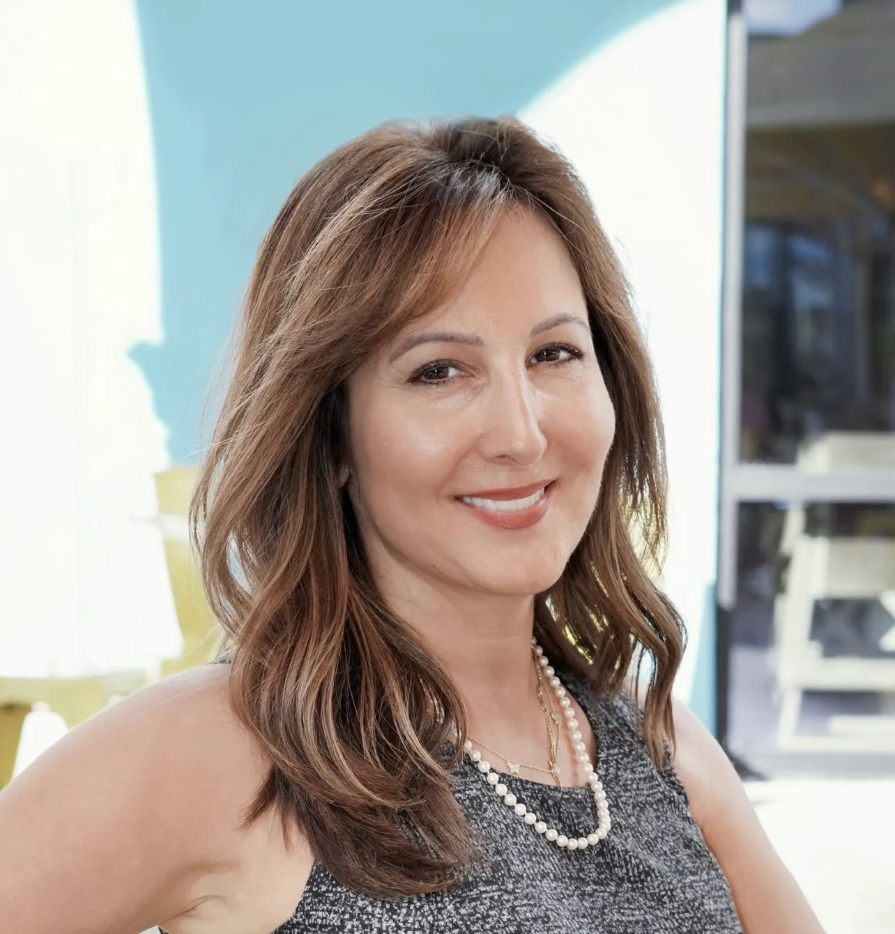 Smiling woman with light brown hair wearing a pearl necklace and sleeveless gray top in bright outdoor setting.