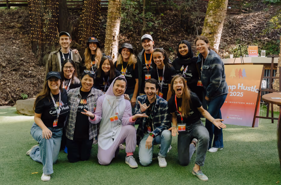 Group of smiling people posing outdoors on grass in front of trees and a Camp Hustle 2025 sign.