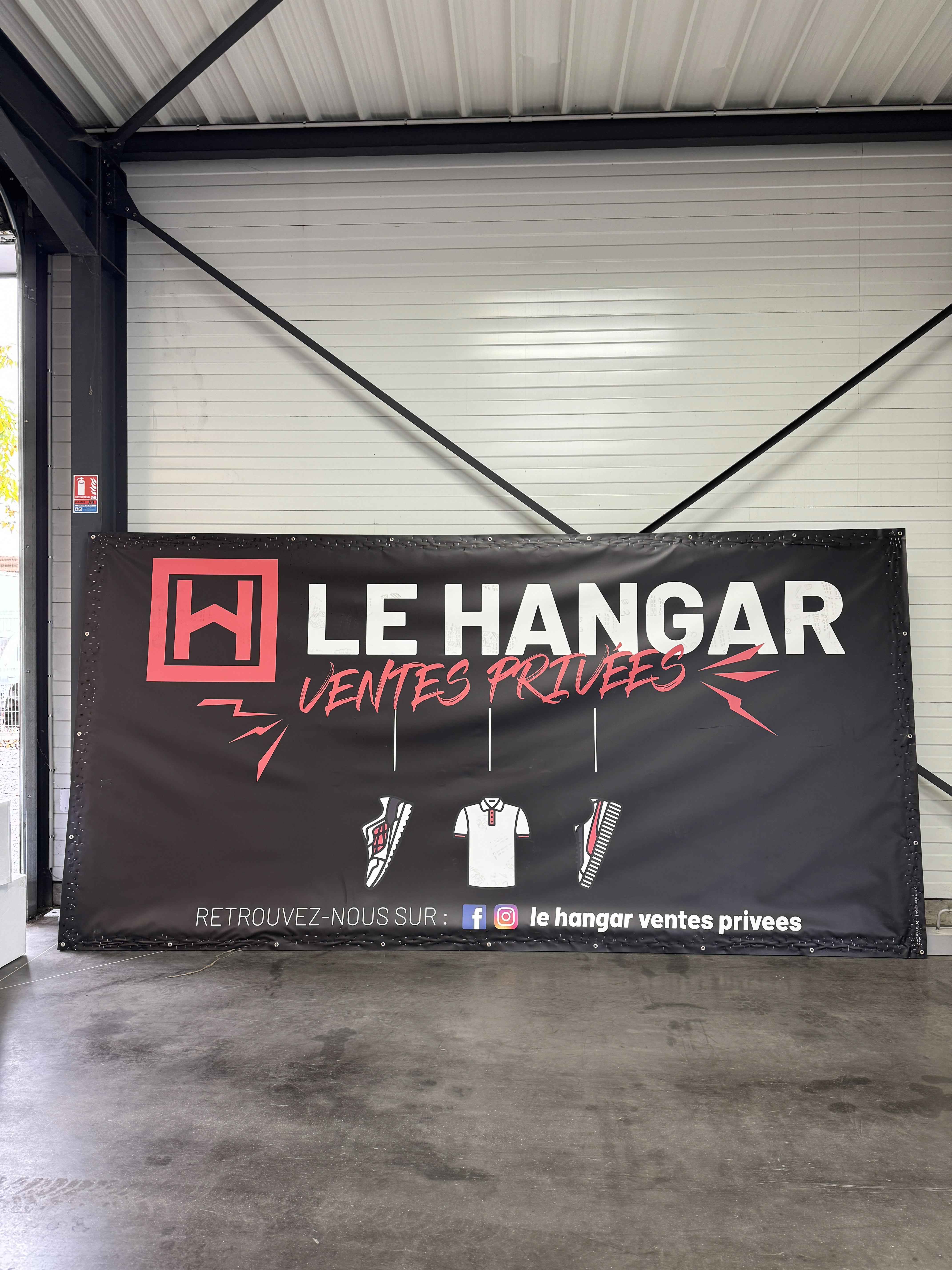 Bâche promotionnelle de “Le Hangar – Ventes Privées” affichée dans un local industriel, montrant le logo rouge et blanc, des illustrations de baskets et vêtements, ainsi que les icônes des réseaux sociaux de la boutique éphémère.