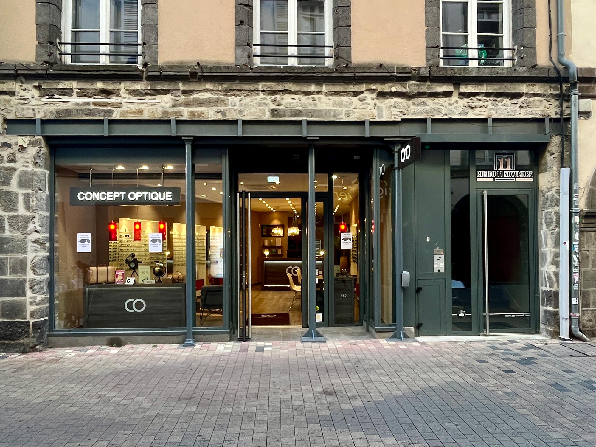 Façade de la boutique “Concept Optique” située au 11 rue du 11 Novembre à Clermont-Ferrand, montrant une vitrine lumineuse avec présentoirs de lunettes et l’entrée principale vitrée de l’enseigne spécialisée en optique.
