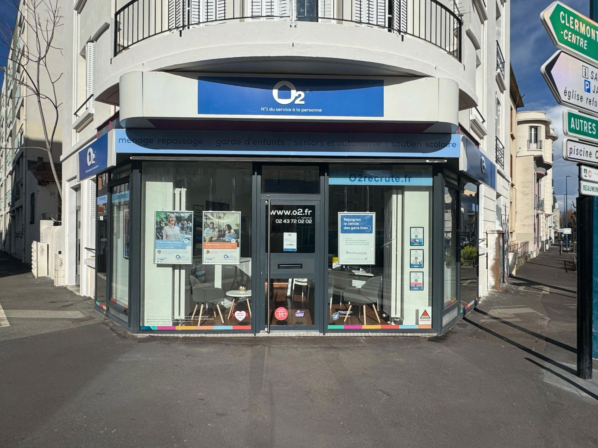 Façade de l’agence O2 à Clermont-Ferrand, avec une devanture vitrée, une enseigne bleue et blanche, et des affiches visibles à l’intérieur. L’agence est située à l’angle d’un immeuble.