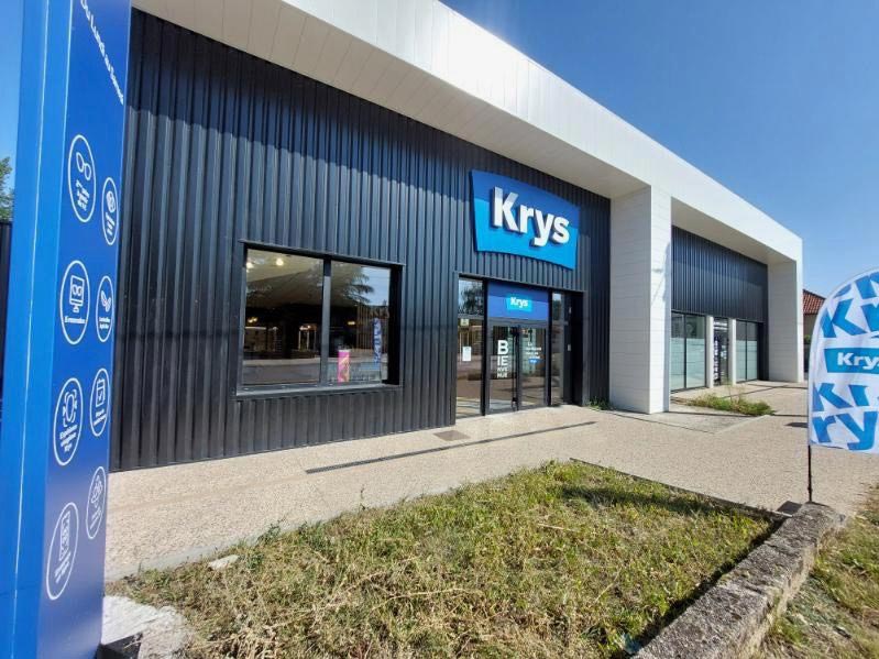 Façade du magasin Krys à Thiers, avec un bâtiment moderne comprenant un bardage gris foncé, une entrée vitrée et un totem bleu indiquant les services de l’enseigne.