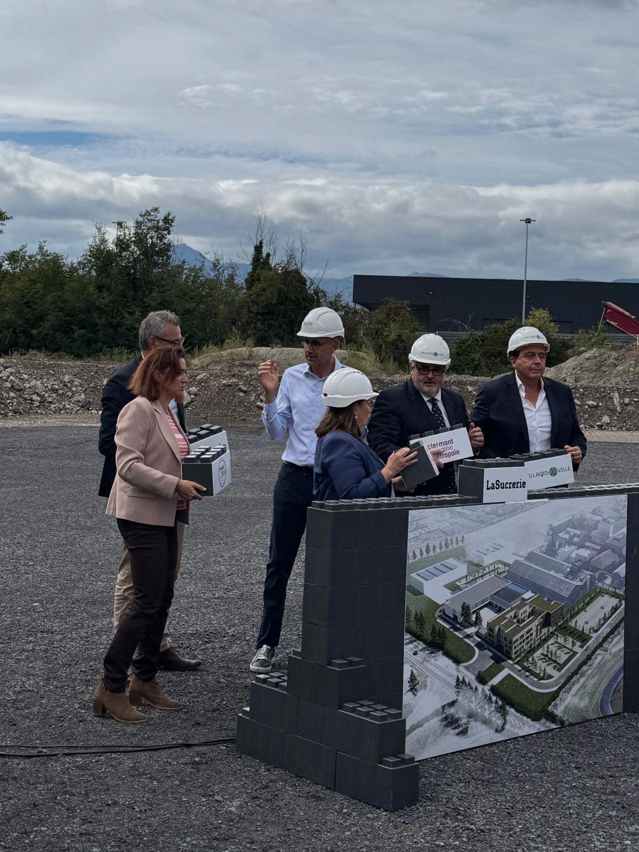 Plusieurs personnes portant des casques de chantier participent à une cérémonie de pose de première pierre sur un terrain en chantier, devant un panneau illustrant le futur bâtiment du site de la Sucrerie de Bourdon.