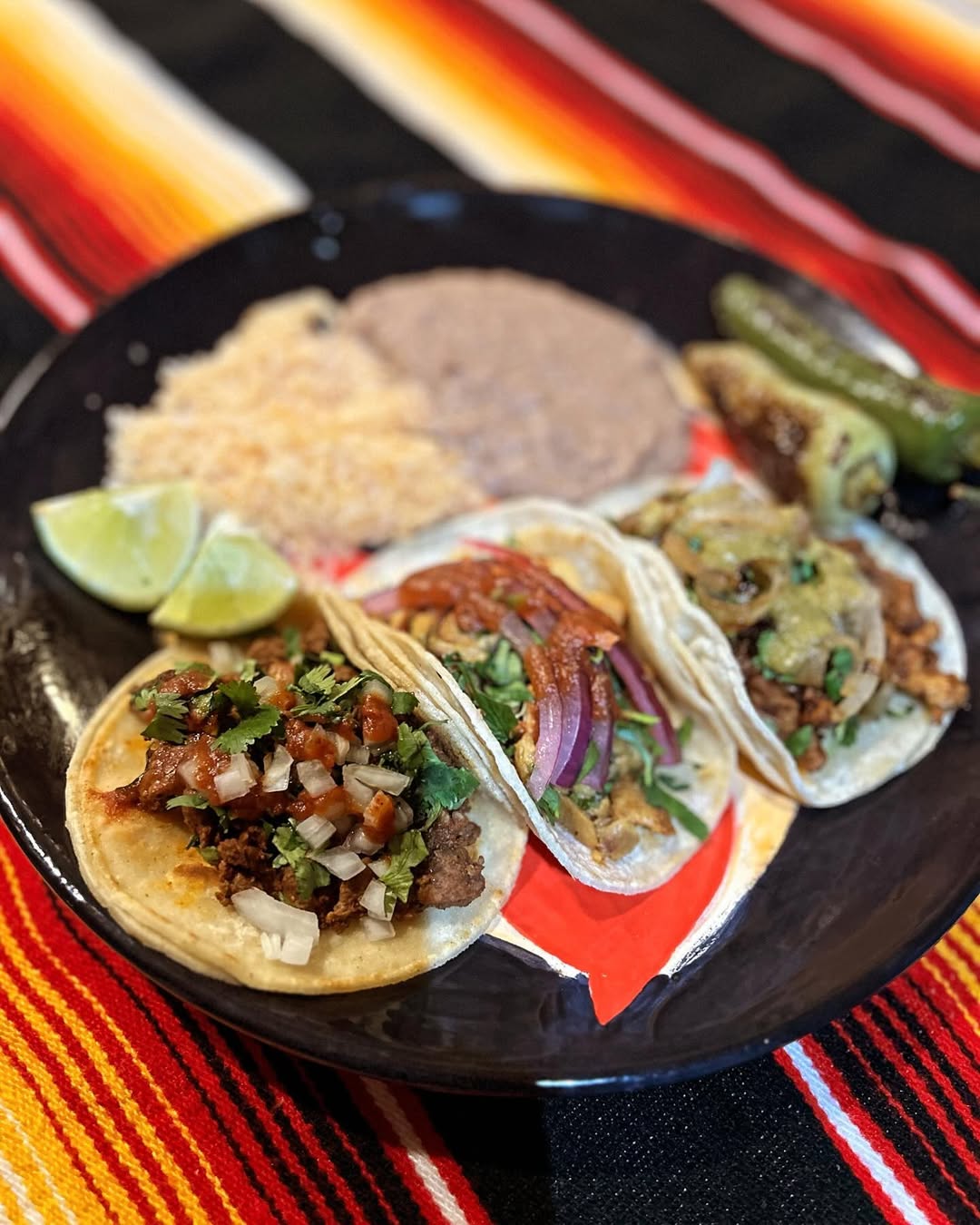 Plate with three tacos topped with onions, cilantro, red sauce, and salsa, served with lime wedges, rice, refried beans, and grilled peppers on a colorful striped tablecloth.