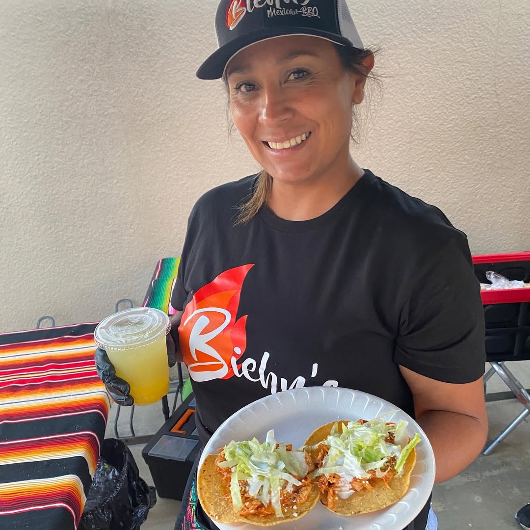 Smiling woman wearing a black hat and t-shirt holding a plate with two tacos topped with lettuce and sauce and a plastic cup with a yellow drink.