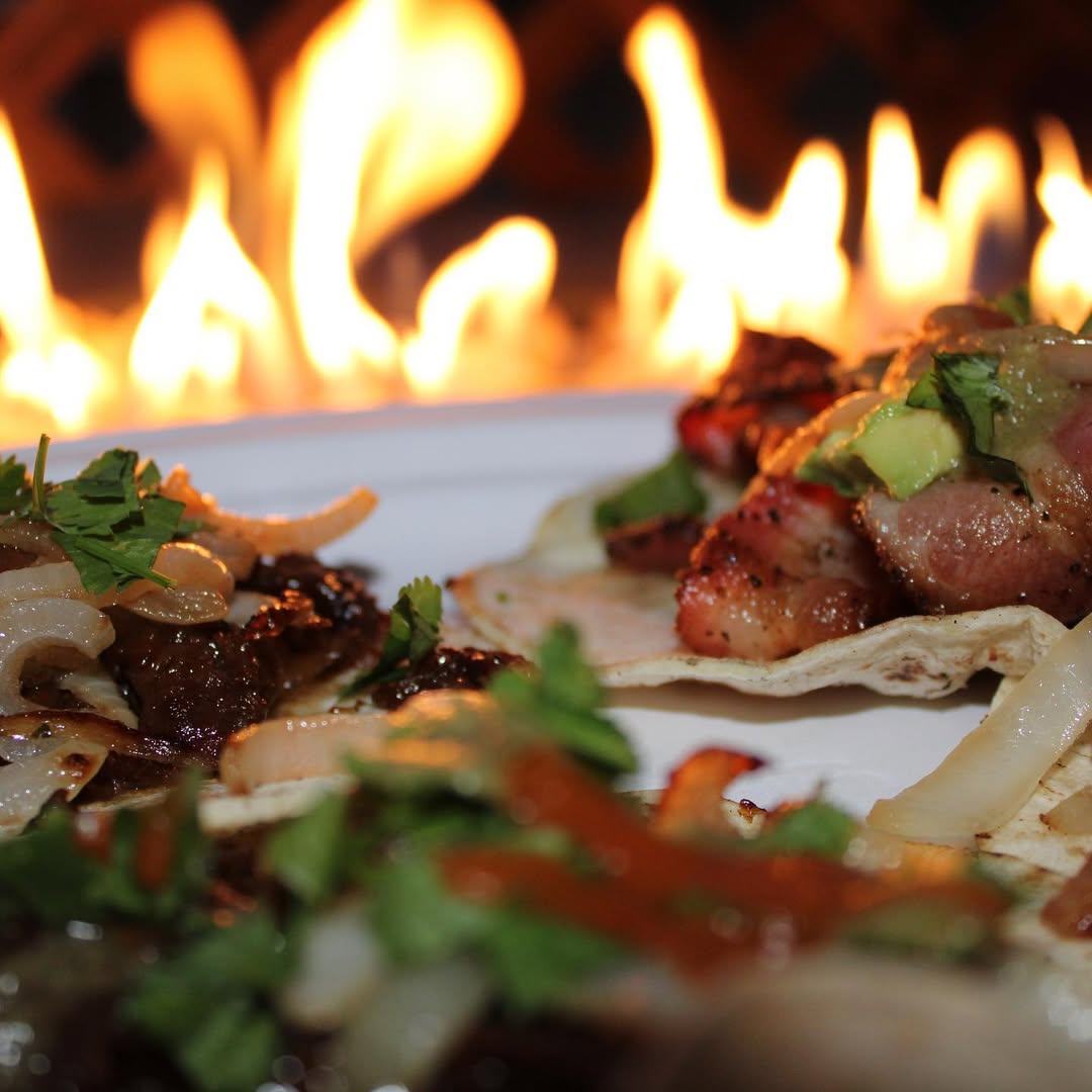 Close-up of three tacos with grilled meat, onions, cilantro, and avocado with flames in the background.