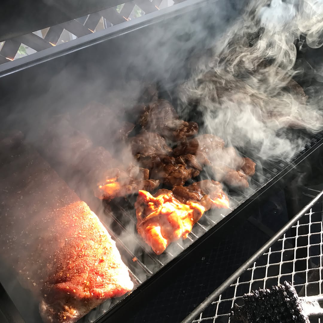 Various cuts of meat smoking on a grill with visible smoke rising around them.