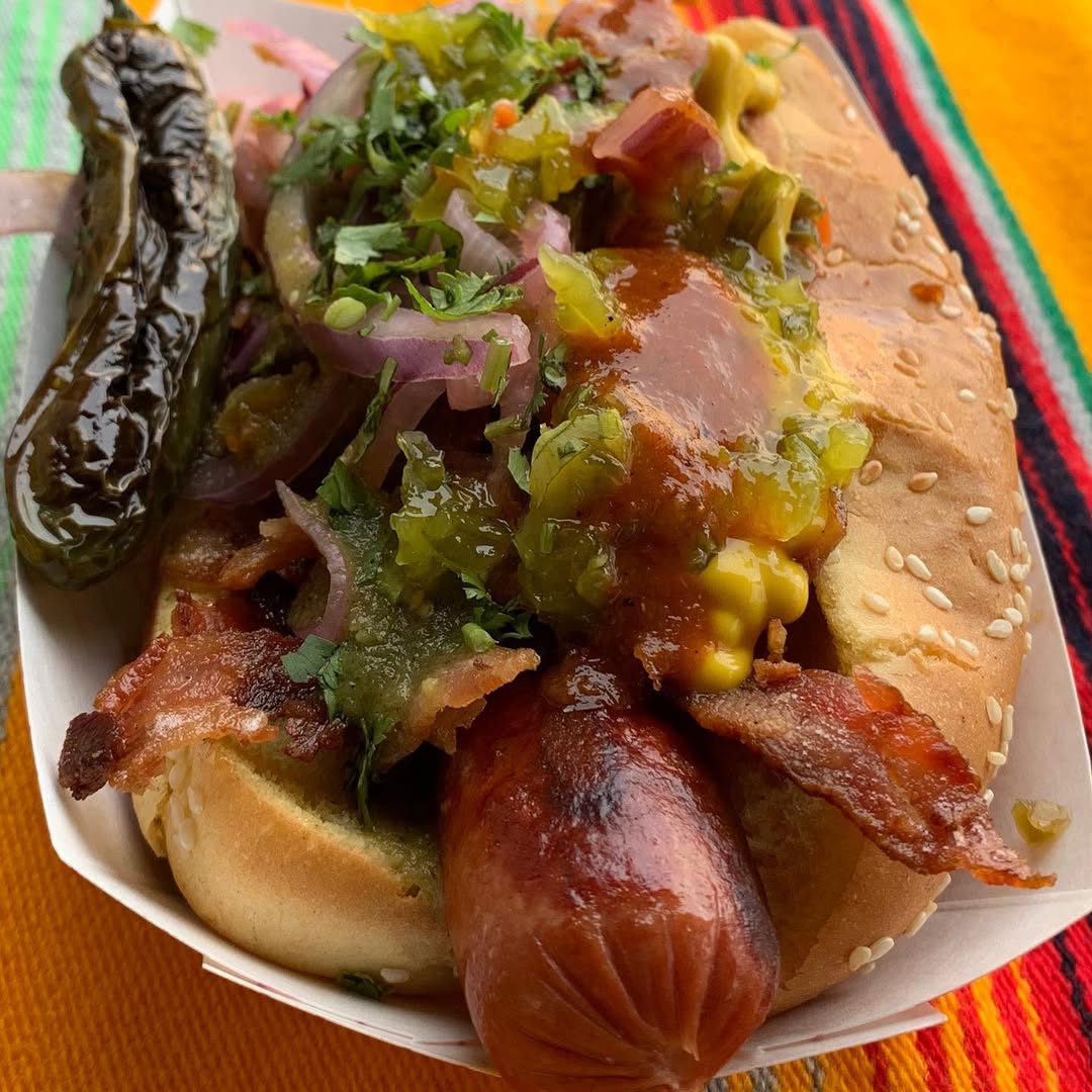 Grilled hot dog topped with bacon, onions, cilantro, pickled relish, mustard, and sauce, served with a pickled pepper on the side in a sesame seed bun.