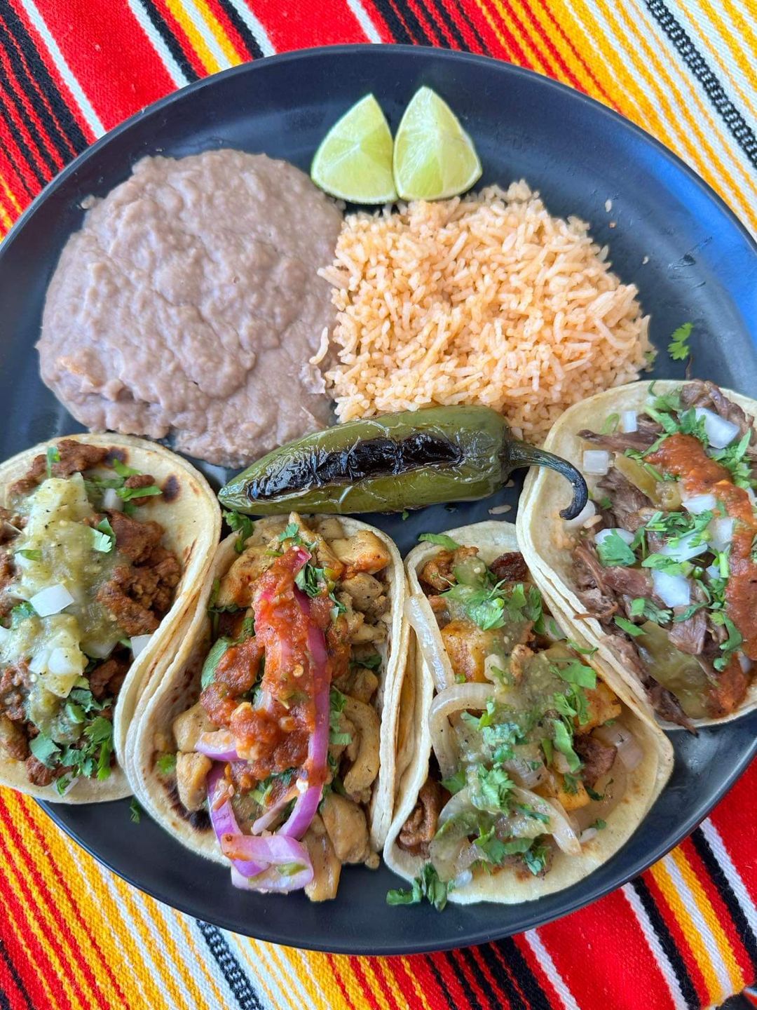 Plate with four tacos topped with salsa, cilantro, and onions, served with refried beans, Mexican rice, two lime wedges, and a grilled jalapeño on a colorful striped tablecloth.