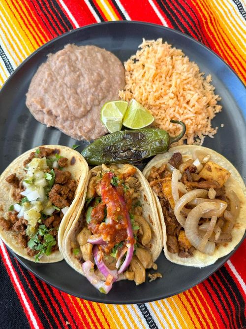 Plate with three assorted tacos topped with onions, cilantro, and salsa, served with refried beans, Mexican rice, a grilled jalapeño, and lime wedges on a colorful striped tablecloth.