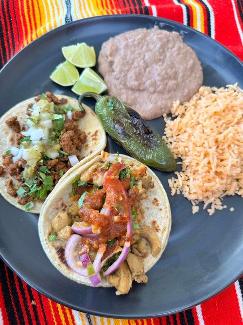 Plate with two tacos topped with meat, onions, salsa, and cilantro, accompanied by lime wedges, refried beans, Mexican rice, and a charred green pepper on a striped tablecloth.