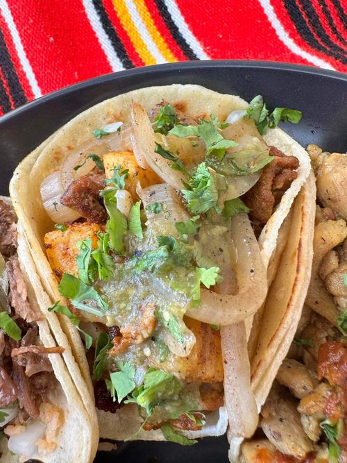 Three street tacos on a black plate with tortillas, grilled meat, onions, cilantro, and green salsa, set on a red striped cloth.