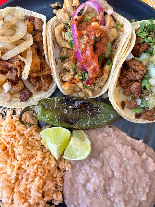 Plate with three tacos filled with grilled meat, chicken with salsa and onions, refried beans, Mexican rice, charred jalapeño, and lime wedges.