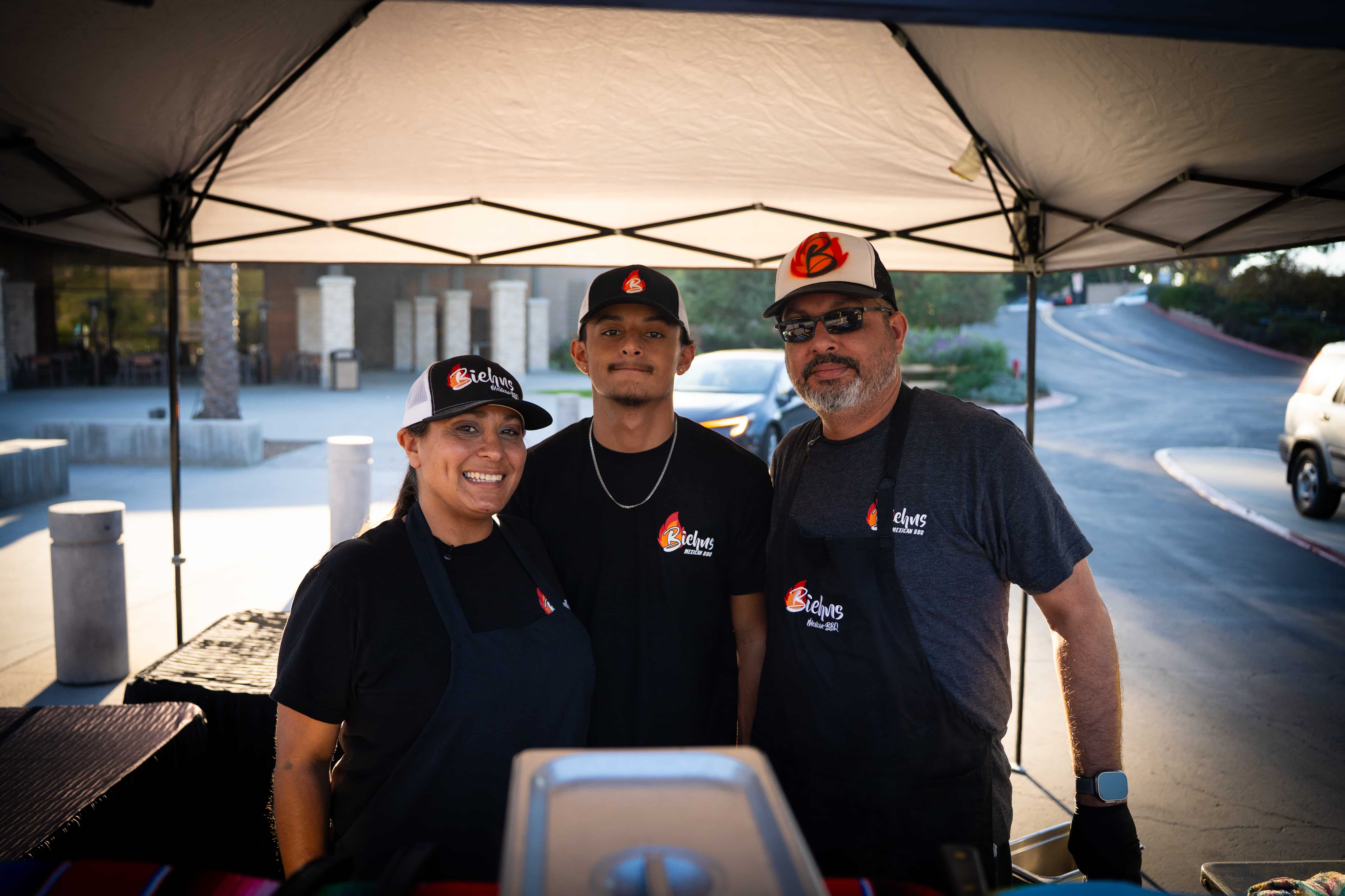 Three people wearing black Biehn's Mexican BBQ shirts and caps standing under a tent at an outdoor event.