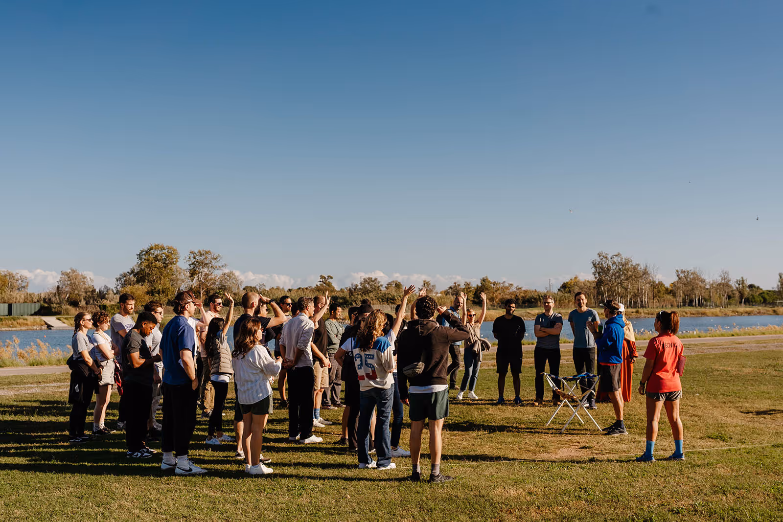 Group of people standing outdoors on grass near a lake on a sunny day, some raising hands during a discussion.