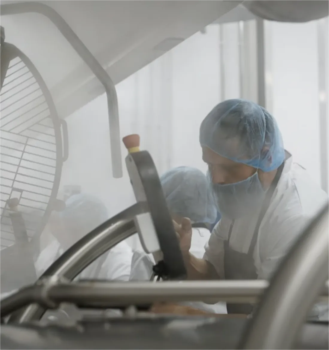 Pracovníci v ochranných síťkách na vlasy a maskách obsluhují průmyslový stroj v potravinářské výrobě.