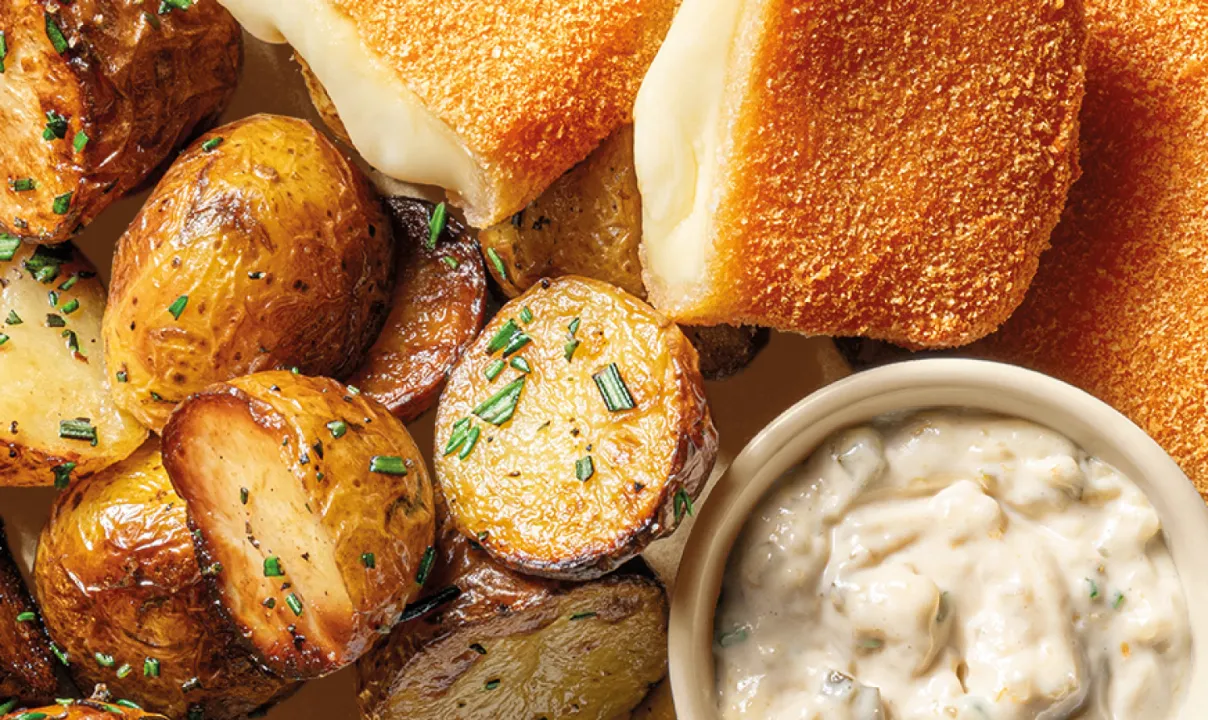 Pečené zlatavé brambory posypané bylinkami vedle kousků smaženého obalovaného sýra a misky smetanového dipu.