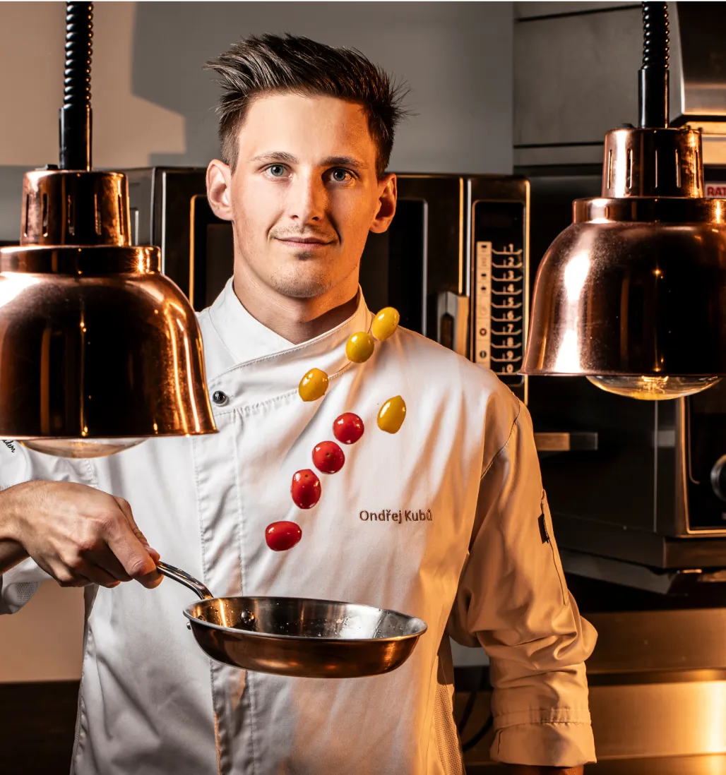 Mladý kuchař v bílé uniformě drží pánev, ve které vesele létají červená a žlutá cherry rajčata.