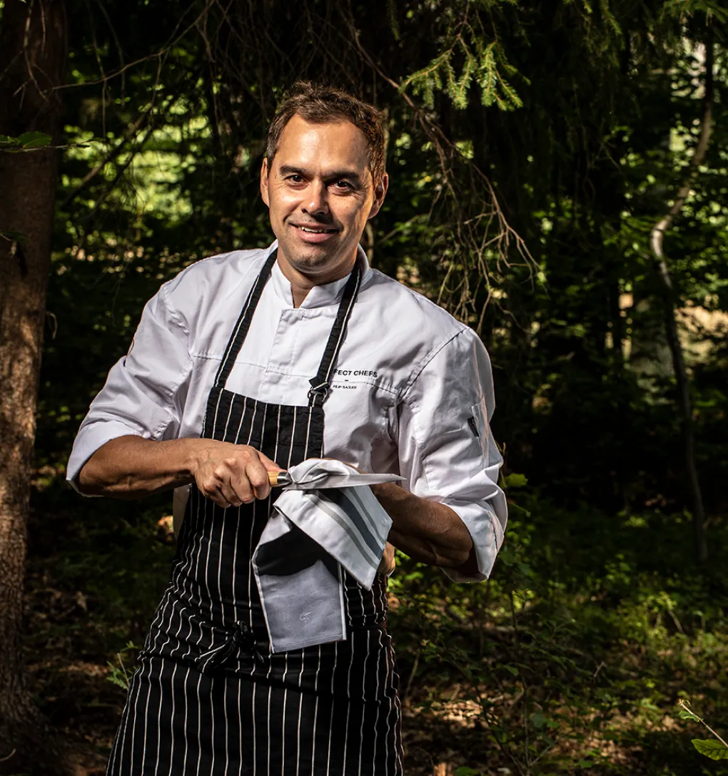 Šéfkuchař s bílou zástěrou a pruhovanou zástěrou utírá nůž v lese.