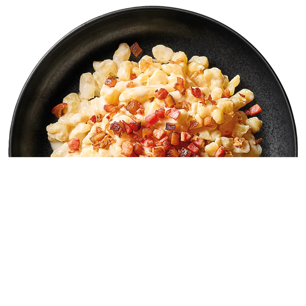 Černý talíř plný halušek s krémovou omáčkou a opečenými kousky slaniny.