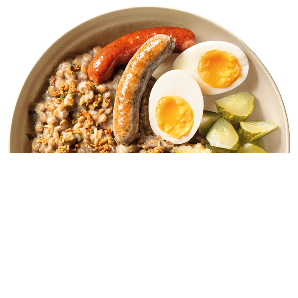 Talíř s čočkovou kaší, dvěma klobásami, vařenými vejci a nakládanými okurkami.