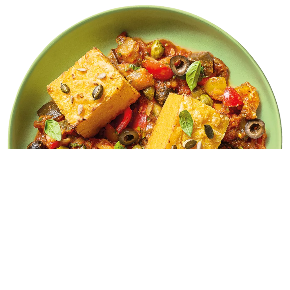 Zelený talíř s caponatou, dušenou zeleninou s olivami, kapary a dvěma kousky polenty posypané semínky a ozdobené bazalkovými lístky.