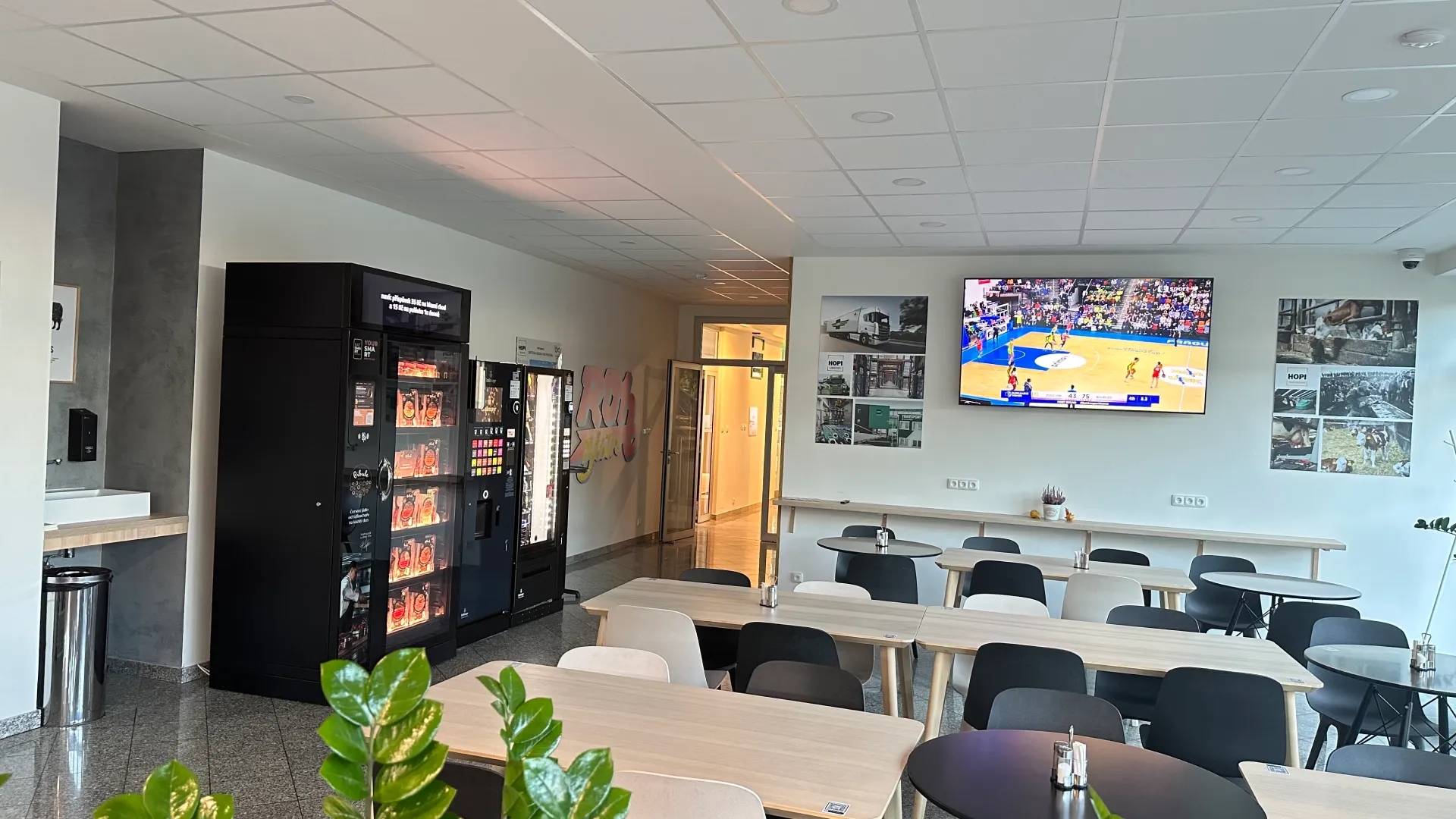 Moderní jídelna s automaty na občerstvení, televizí na stěně a stolky s židlemi v přední části.