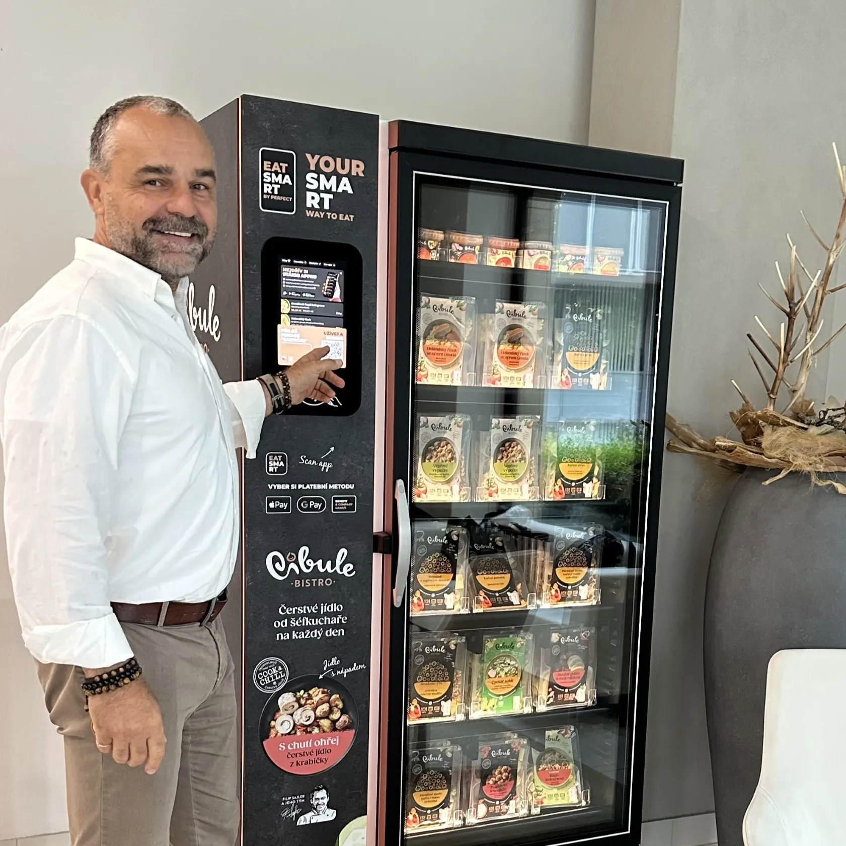 Muž v bílé košili stojí u prodejního automatu s čerstvým jídlem značky Cibule Bistro a volí na dotykové obrazovce možnost platby.