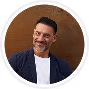 Smiling middle-aged man with dark hair and beard, wearing a dark blazer over a white shirt, against a brown background.