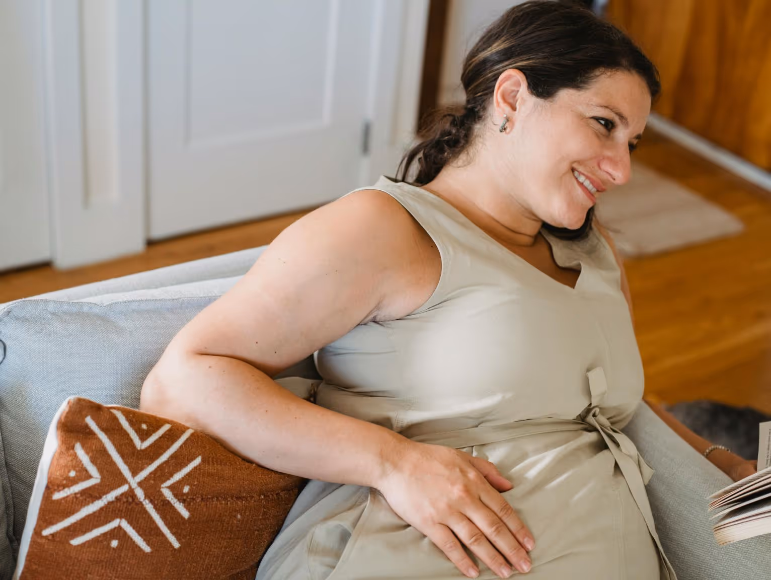 Smiling pregnant woman wearing beige dress reclining on a grey couch talking to therapist about postpartum concerns.