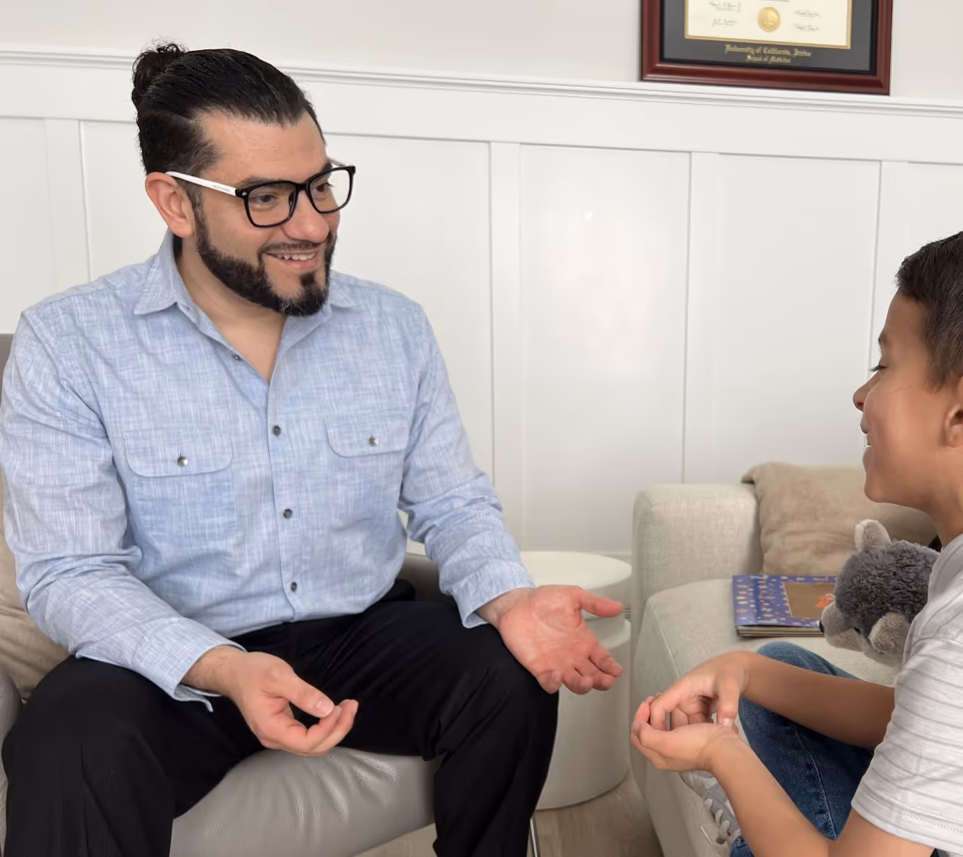 Smiling man with glasses evaluating a young boy holding a stuffed animal in a cozy living room.
