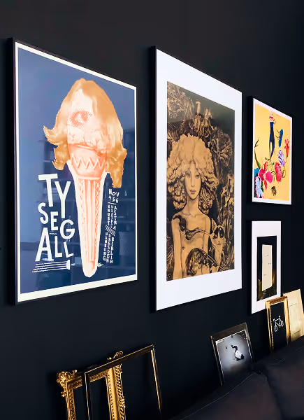 Gallery wall with various framed artworks, including a surreal portrait with wavy hair, a colorful abstract piece, and typographic poster featuring an ice cream cone and words.