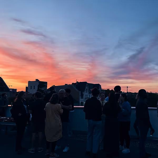 Group of people socializing on a rooftop at sunset with colorful sky and silhouetted buildings.