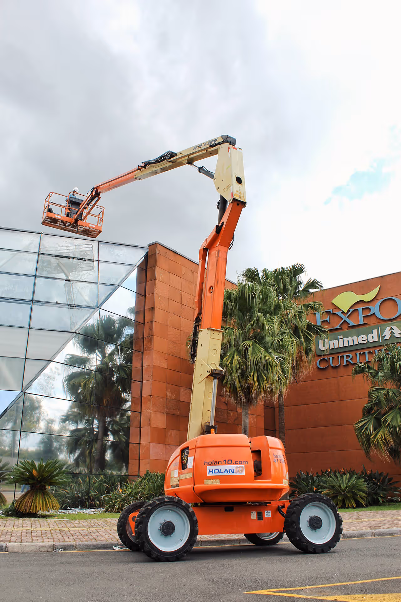 Elevador articulado laranja com trabalhador em plataforma próxima à fachada de vidro e tijolos de um edifício com palmeiras.