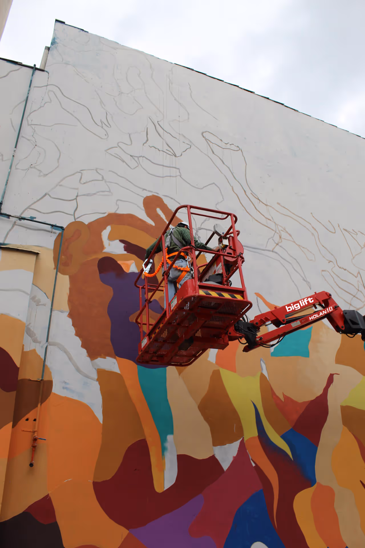 Artista em um elevador aéreo pintando um mural colorido abstrato em uma grande parede externa.