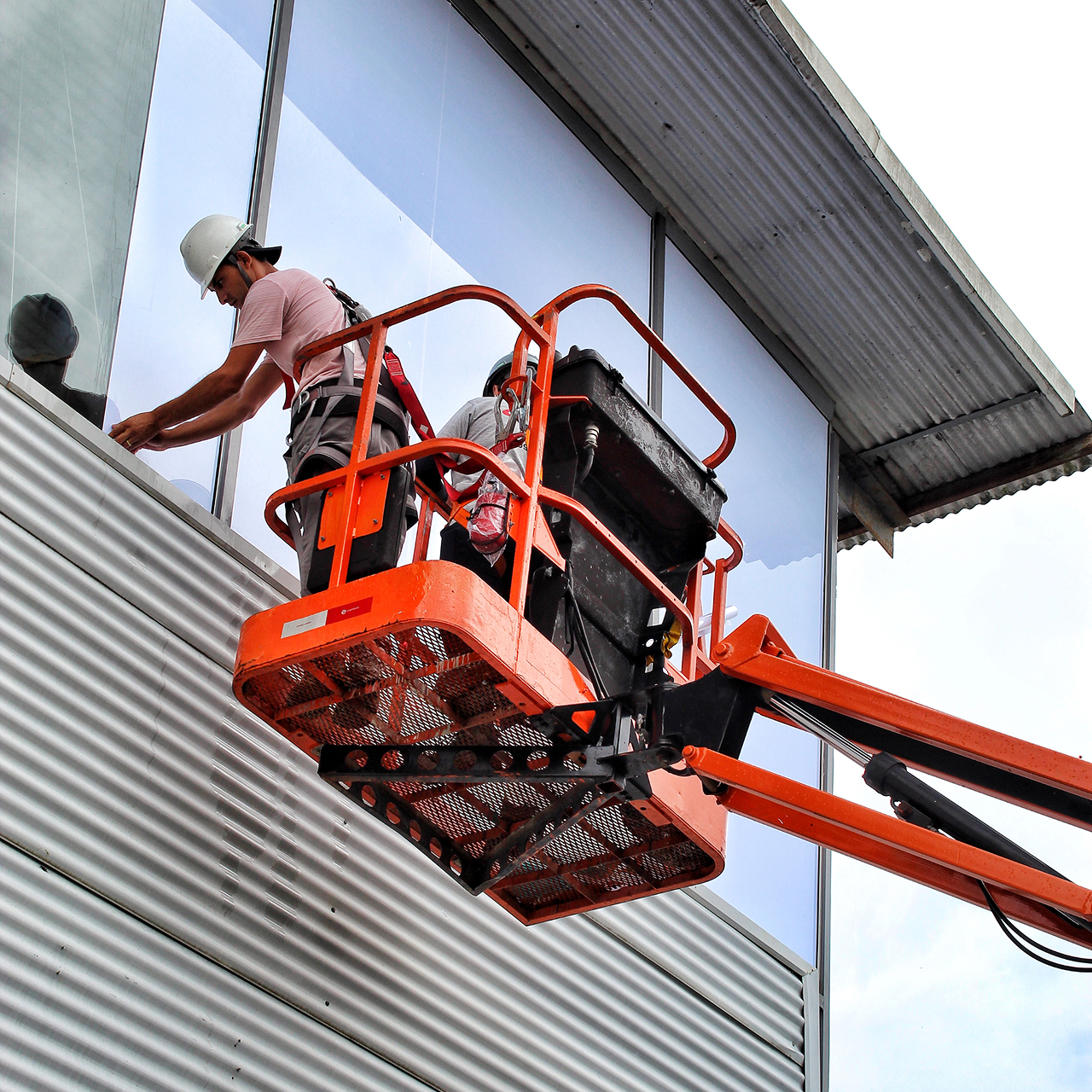 Dois trabalhadores com capacetes em uma plataforma elevada laranja consertando uma janela de vidro em um prédio metálico.
