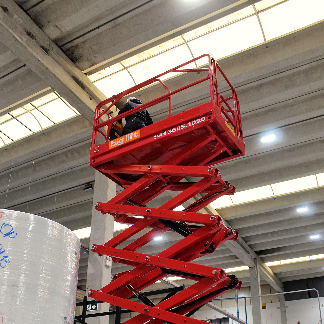 Trabalhador em plataforma elevatória vermelha em um armazém industrial com teto de concreto.