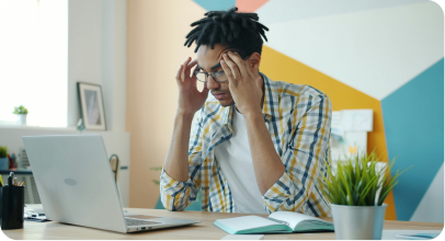 Young man wearing glasses and a checkered shirt sitting at a desk with a laptop, holding his head in frustration or stress.