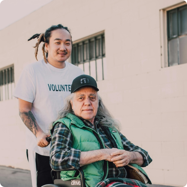 Young volunteer with dreadlocks standing behind an older man in a wheelchair outside a building.