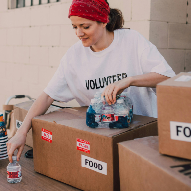 Volunteer in a red bandana packing bottled water and food boxes labeled 'FOOD' on a table.