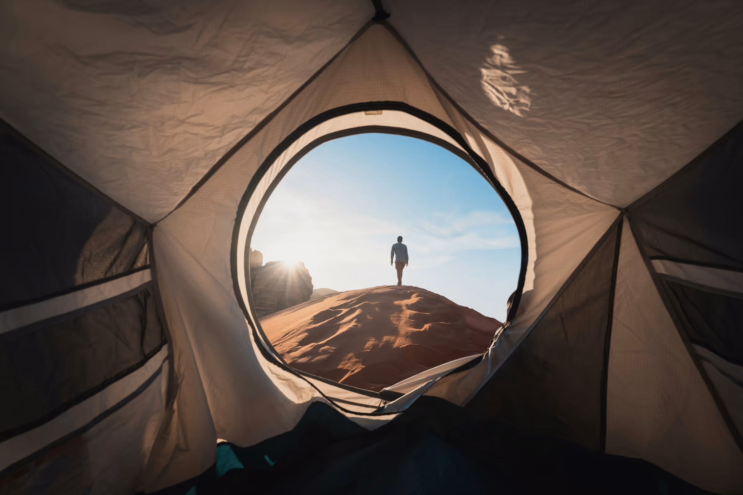 Person standing on a sunlit sand dune seen through the circular opening of a tent.