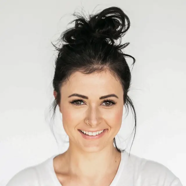 Smiling woman with dark hair in a messy bun wearing a white shirt against a plain background.