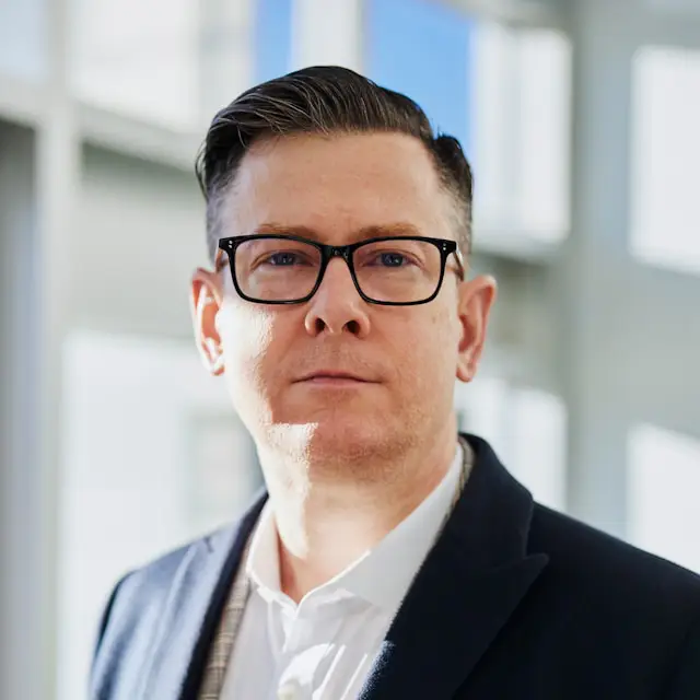 Man with short dark hair and glasses wearing a suit jacket and white shirt in a bright modern office.