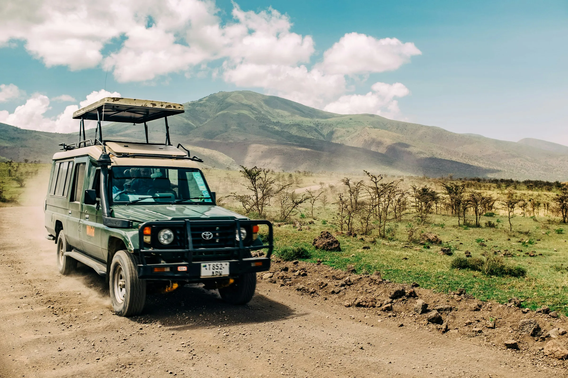 Green safari jeep driving on a dirt road with mountains and scattered trees in the background under a partly cloudy sky.