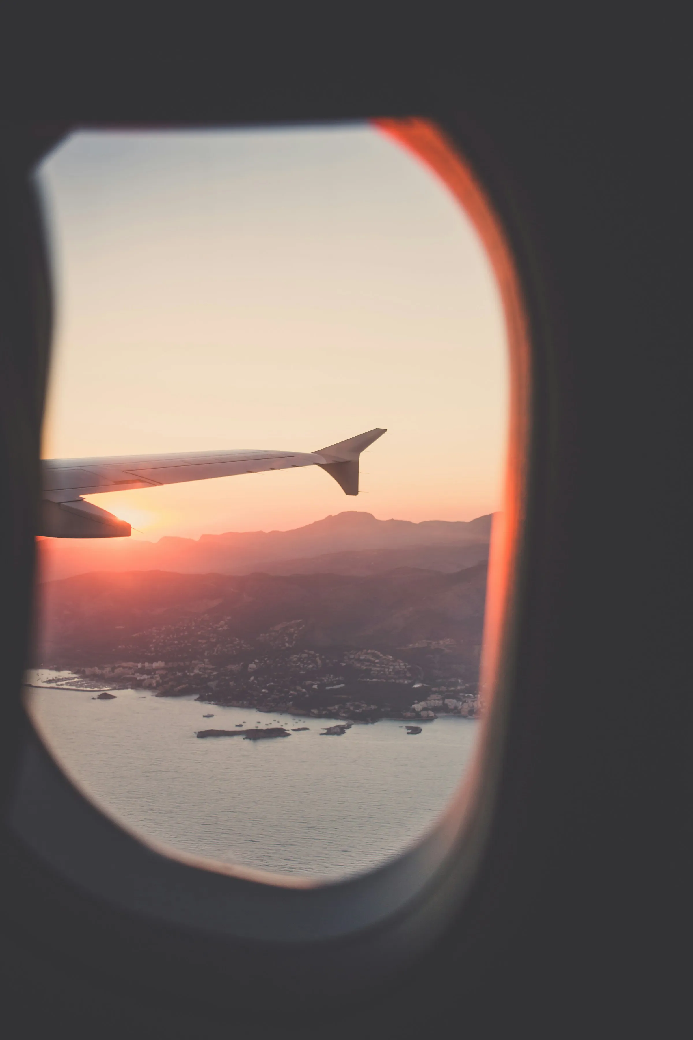 View of a coastal landscape with mountains at sunset through an airplane window showing the wing and sun's orange glow.