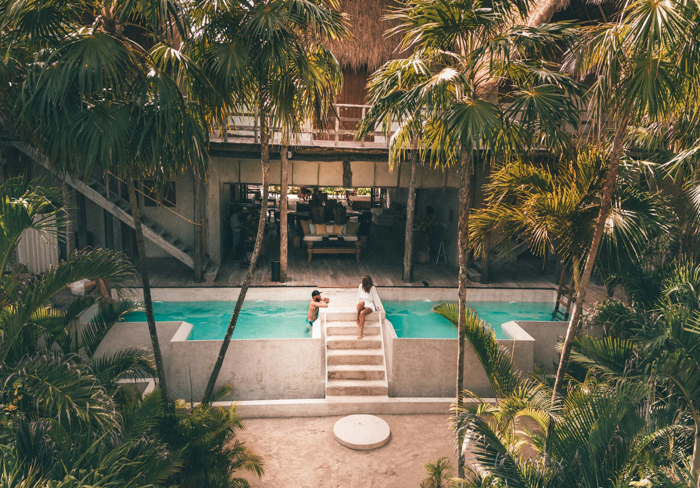 Two people relaxing near a turquoise pool surrounded by tropical palm trees and a rustic building with thatched roofing.