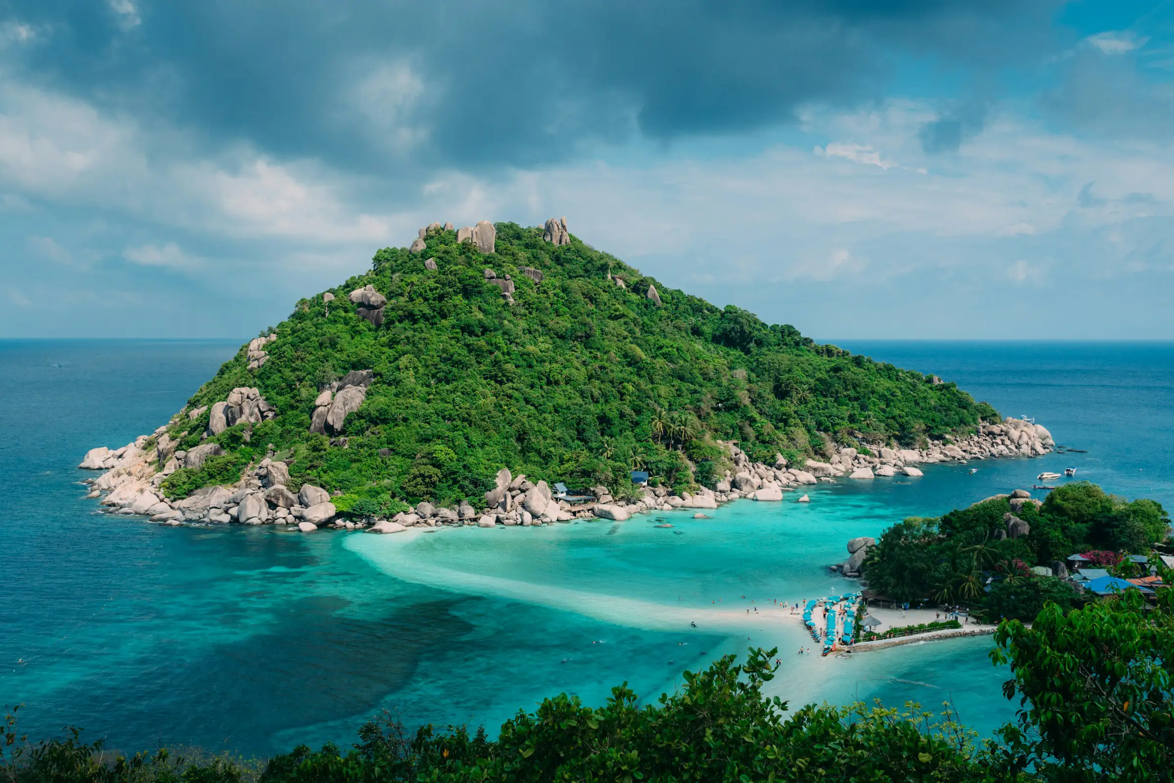 Lush green island surrounded by turquoise and dark blue ocean waters under a partly cloudy sky.
