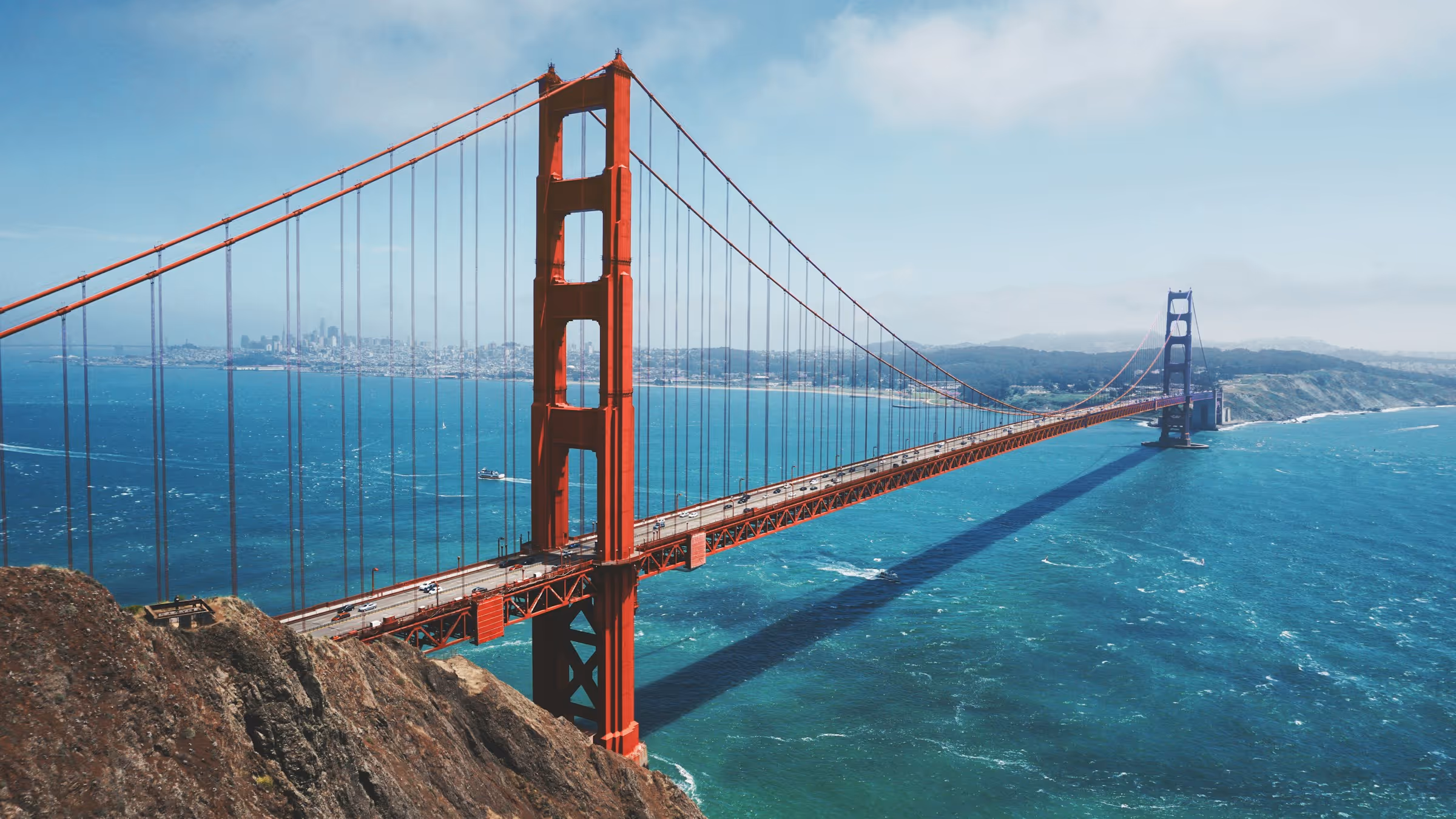 Golden Gate Bridge spanning across blue water with city and hills in the background on a clear day.